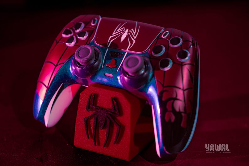 May include: A custom-painted video game controller with a Spider-Man theme. The controller is primarily red with black spiderweb detailing and a white spider emblem. The design includes blue and purple accents. A red stand with a spider logo supports the controller.
