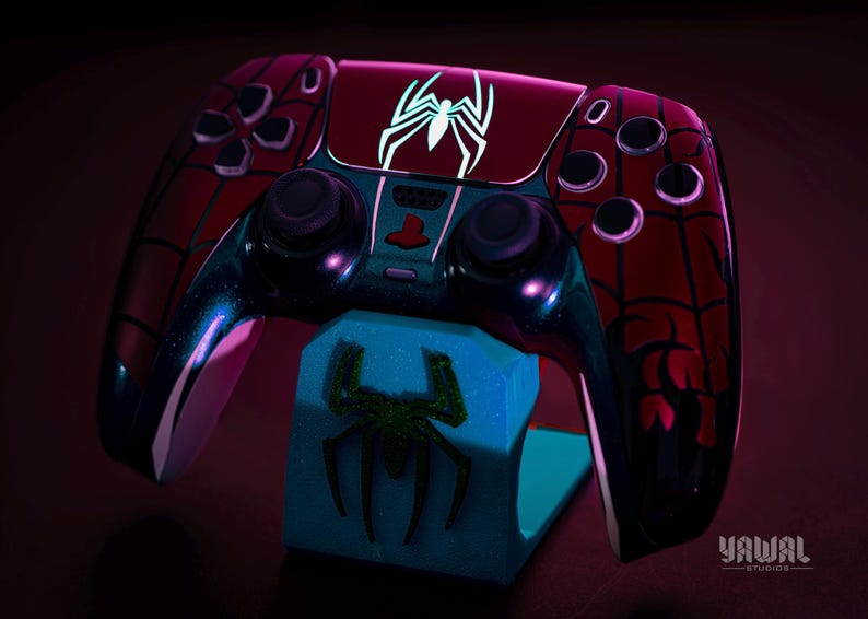 May include: A custom-designed video game controller in a Spider-Man theme. The controller is primarily red with black web patterns and a white spider emblem. It rests on a blue stand with a green spider design.