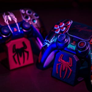 May include: Two custom-painted PlayStation 5 controllers with a Spider-Man theme. The controllers feature red, black, and blue accents, with a spider logo and web design. Each controller rests on a matching stand.