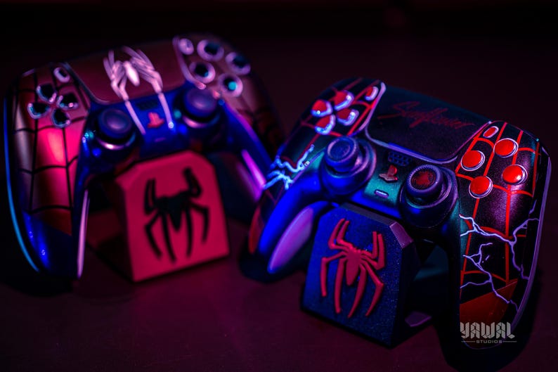 May include: Two custom-painted PlayStation 5 controllers with a Spider-Man theme. The controllers feature red, black, and blue accents, with a spider logo and web design. Each controller rests on a matching stand.