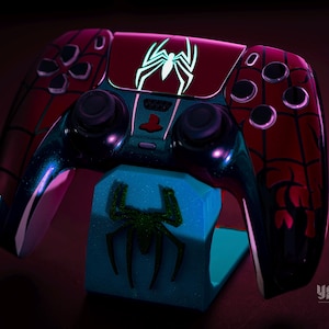 May include: A custom-designed video game controller in a Spider-Man theme. The controller is primarily red with black web patterns and a white spider emblem. It rests on a blue stand with a green spider design.