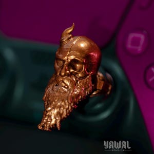 May include: A gold-colored controller accessory shaped like a bearded skull. The skull has a prominent brow, a long beard, and a horn-like protrusion. The accessory is set against a dark green and purple background, with the text "YAWAL STUDIOS" visible.