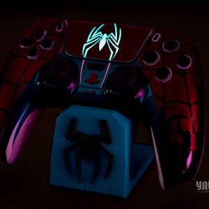 May include: A custom-designed video game controller with a Spider-Man theme, featuring a red and black color scheme with a glowing spider emblem. The controller rests on a blue stand with a matching spider logo. The image is set against a dark background.