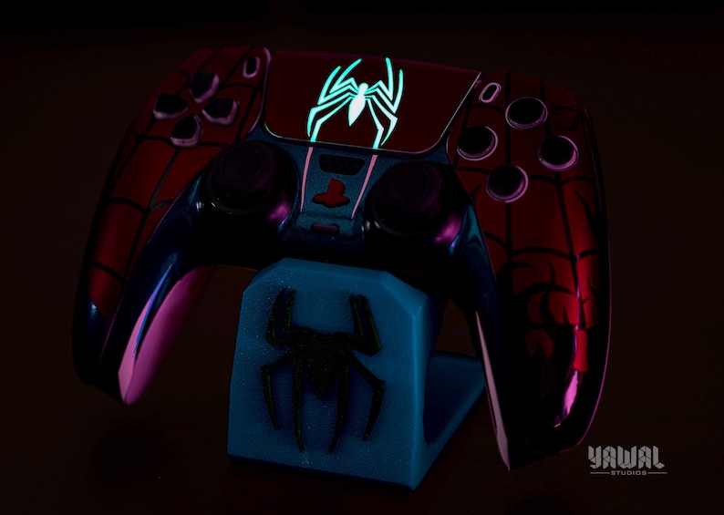 May include: A custom-designed video game controller with a Spider-Man theme, featuring a red and black color scheme with a glowing spider emblem. The controller rests on a blue stand with a matching spider logo. The image is set against a dark background.