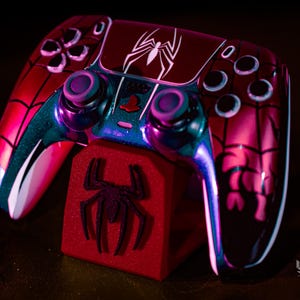May include: A custom-designed video game controller with a Spider-Man theme. The controller is primarily red with black accents and a white spider logo. It rests on a red stand with a spider emblem.