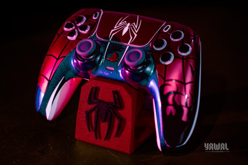May include: A custom-designed video game controller with a Spider-Man theme. The controller is primarily red with black accents and a white spider logo. It rests on a red stand with a spider emblem.