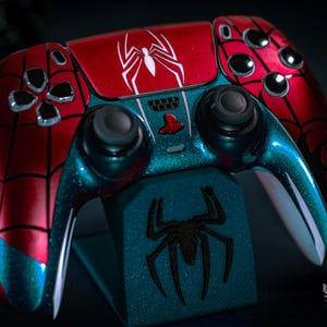May include: A custom-painted video game controller in red and teal, featuring a spiderweb design and a white spider emblem. The controller rests on a matching stand with a black spider graphic. The controller has black joysticks and buttons.