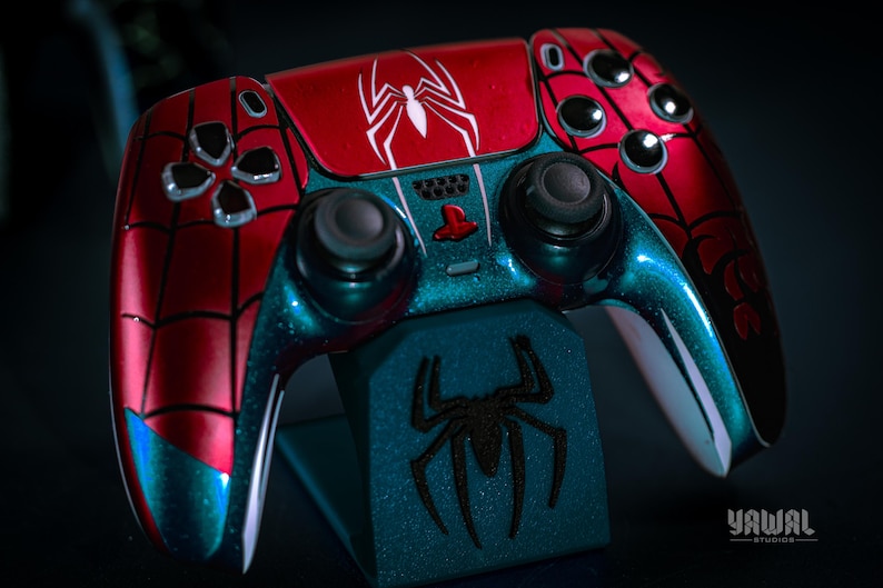May include: A custom-painted video game controller in red and teal, featuring a spiderweb design and a white spider emblem. The controller rests on a matching stand with a black spider graphic. The controller has black joysticks and buttons.