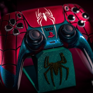 May include: A custom-painted PlayStation 5 controller in a Spider-Man theme. The controller features a red and blue color scheme with a spiderweb design and a white spider emblem. It rests on a matching green stand with a spider logo.