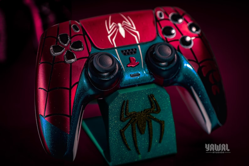 May include: A custom-painted PlayStation 5 controller in a Spider-Man theme. The controller features a red and blue color scheme with a spiderweb design and a white spider emblem. It rests on a matching green stand with a spider logo.