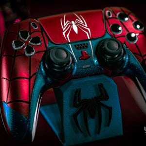 May include: A custom-painted Spider-Man-themed video game controller. The controller features a red and blue color scheme with a white spider emblem on the top. The controller is resting on a blue stand with a black spider logo.