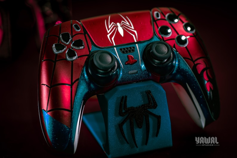 May include: A custom-painted Spider-Man-themed video game controller. The controller features a red and blue color scheme with a white spider emblem on the top. The controller is resting on a blue stand with a black spider logo.