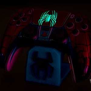 May include: A red and black Spider-Man-themed game controller with a glowing green spider emblem. The controller rests on a blue stand with a matching spider design. The design glows in the dark.