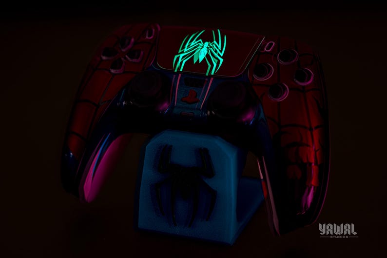 May include: A red and black Spider-Man-themed game controller with a glowing green spider emblem. The controller rests on a blue stand with a matching spider design. The design glows in the dark.