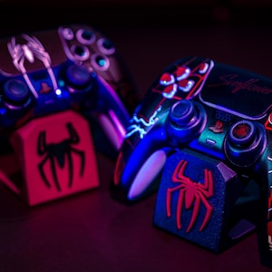 May include: Two custom-painted game controllers with matching stands. The controllers feature a Spider-Man theme with red, black, and blue accents. One controller has the text "Skyflower". The stands display a red spider logo.