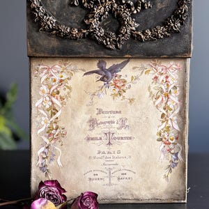 May include: A rectangular wall-mounted box featuring a black top panel with ornate bronze-colored floral molding. The front panel displays a vintage-style print with a bird, floral garlands, and French text including "Peinture" and "Paris".