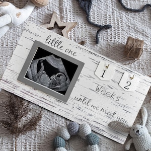 May include: A baby announcement display with a framed sonogram, a knitted sweater, and a plush giraffe. The wooden sign reads "little one" and "12 Weeks until we meet you". Other items include a knitted hat and a stuffed bunny.