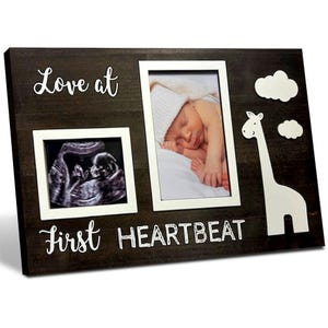 May include: A dark brown wooden picture frame with two photo openings and white text that reads "Love at First HEARTBEAT." One photo opening contains a baby ultrasound, and the other holds a photo of a sleeping baby. A white giraffe and clouds are on the right.