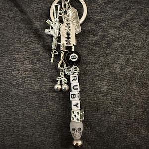 May include: Silver keychain with various charms, including a miniature rifle, a razor blade, an 8-ball, and dice. The keychain also features a series of lettered beads spelling "RUBY", and a skull charm. The background is a dark gray fabric.