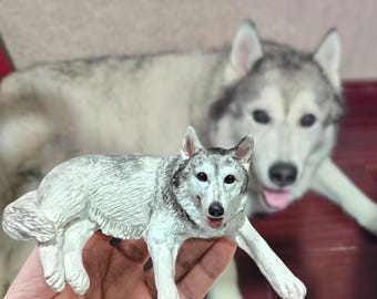 Puede incluir: Una figurita de lobo gris y blanco acostada de lado con las patas extendidas. La figurita está hecha de plástico y tiene un aspecto realista.