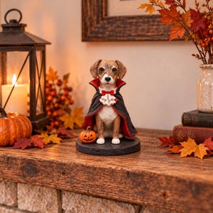 Peut inclure: Une figurine de chien marron et blanc portant une cape noire à doublure rouge, un col à volants blanc et un nœud papillon rouge. Le chien est assis sur un socle rond noir à côté d'une petite citrouille orange avec un visage sculpté.