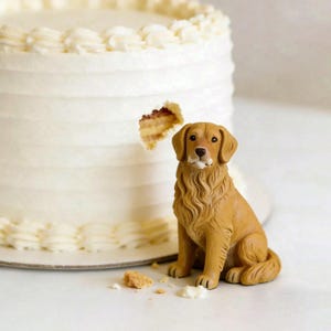 May include: A golden retriever figurine sits beside a white frosted cake with a bite taken out. The cake has a simple design with white frosting and a decorative border. Cake crumbs and frosting are scattered around.