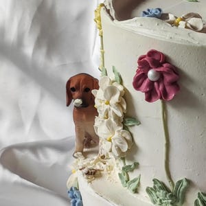 May include: A white cake decorated with buttercream frosting and fondant flowers. The cake features a small dog figurine and a variety of flowers, including white, pink, and blue.