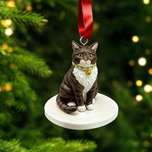 May include: A Christmas tree ornament shaped like a brown and white tabby cat. The cat is sitting on a white disc and wearing a collar with a small gold bell. A red ribbon is attached for hanging.