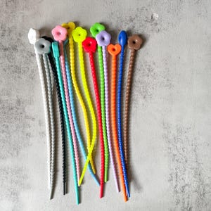 Puede incluir: Una colección de cierres de cordones de silicona de colores, con forma de flor. Los cierres son de varios colores, incluyendo rojo, naranja, amarillo, verde, azul, morado, rosa y blanco.