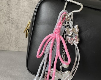 Puede incluir: Un llavero rosa, gris y plateado con dijes de lazo y un mosquetón. El llavero está sujeto a un bolso negro. Los dijes de lazo son plateados y tienen un diseño delicado. Los cordones rosas y grises están anudados.