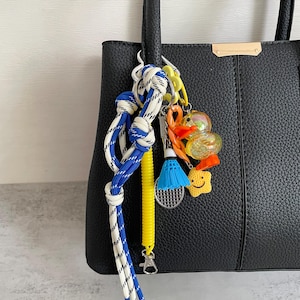 Può includere: Una borsa tote in pelle nera con una varietà di portachiavi colorati. I portachiavi includono un design a corda blu e bianca, un volano da badminton giallo e vari ciondoli originali, tra cui una stella sorridente e ciondoli a forma di anatra.
