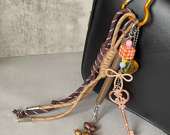 Könnte beinhalten: Ein dekorativer Schlüsselanhänger mit einem goldenen Karabinerhaken, der an einer schwarzen Tasche befestigt ist. Der Schlüsselanhänger verfügt über einen Ballonhund-Anhänger, einen Schlüsselanhänger, Perlen und geflochtene Kordeln in Braun- und Beigetönen.