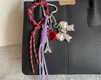 May include: A black leather handbag with a silver charm attached. The charm features a red heart, a silver Eiffel Tower, and a silver bow. The charm is attached to the bag with a red, white and purple rope.