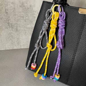 Op de afbeelding: Drie kleurrijke touw sleutelhangers hangen aan een zwarte leren tas. De sleutelhangers zijn grijs, geel en paars met decoratieve kralen aan de onderkant. De tas heeft een goudkleurige sluiting.