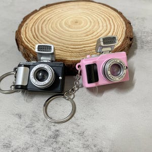 Op de afbeelding: Twee miniatuur camera sleutelhangers, een zwart en een roze, met zilveren accenten en een flitser. Beide camera's hebben een lens met de tekst "ZHAOXIANGJI OPTICAL 1:3.5 F=35mm" rond de lens.