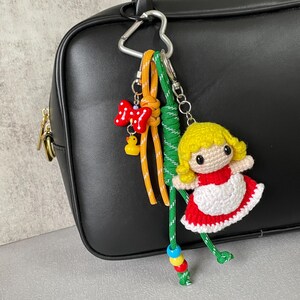 Op de afbeelding: Een handgemaakte sleutelhanger met een gehaakte pop in een rode jurk met een witte schort, geel haar en een schattig gezicht. De sleutelhanger bevat ook een rode strik, een gele eendenbedel en kleurrijke kralen.
