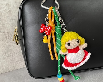 May include: A handmade keychain featuring a crocheted doll in a red dress with a white apron, yellow hair, and a cute face. The keychain also includes a red bow, a yellow duck charm, and colorful beads.