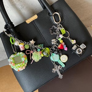 May include: A black textured handbag adorned with a multi-charm silver-tone chain. The charms include a green floral-encrusted digital pet, a miniature camera, a key, a cross, a smiley face, a strawberry, a popcorn bucket, and various colourful beads.