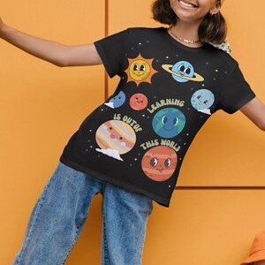 Pode incluir: T-shirt preto com um design colorido do sistema solar. Apresenta planetas sorridentes e o texto "Learning is out of this world." O gráfico inclui um sol sorridente, Saturno, Terra e outros planetas. A camisa é feita de um material macio.