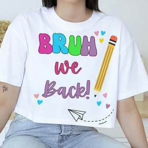 May include: White cropped t-shirt with a colorful "BRUH WE BACK!" graphic and a yellow pencil with a pink eraser. The shirt has a small paper airplane graphic and pink, blue, and red hearts.