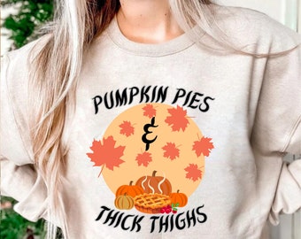 Könnte beinhalten: Ein beiger Pullover mit einem Grafikdesign, das eine Kürbispastete, Kürbisse und Herbstblätter zeigt. Der Text auf dem Pullover lautet "Pumpkin Pies & Thick Thighs".