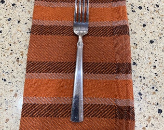 May include: A silver fork placed on top of a folded orange and brown striped napkin.