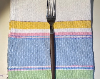 May include: A stainless steel fork with a dark brown handle rests on a white woven napkin. The napkin features horizontal stripes in yellow, pink, blue, green, and purple. The napkin is a kitchen or dining accessory.