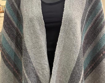 May include: A gray woven poncho with stripes of teal, purple, and gray. The poncho has a fringed hem and is displayed on a mannequin. The overall style is bohemian and suitable for fall or winter wear.