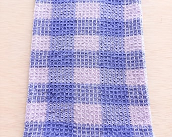 May include: A folded blue and lavender checkered kitchen towel. The towel has a waffle weave texture and is made of a soft fabric. The checkered pattern is made up of alternating squares of blue and lavender.