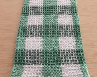 May include: A folded green, white, and dark green checkered kitchen towel. The towel has a waffle weave texture and is made of a soft, absorbent material. The checkered pattern is a classic design, suitable for any kitchen.