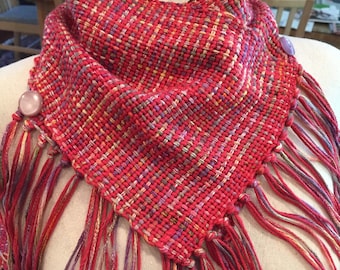 May include: A red, pink, and cream woven scarf with fringe. The scarf has a geometric pattern and is decorated with two small purple buttons.