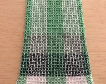 May include: A folded green and white waffle weave kitchen towel. The towel features a gradient design, transitioning from white to light green, then dark green, and back to white. The towel is made of a textured fabric.