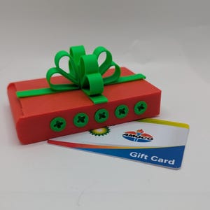 May include: A red gift box with a green bow and green accents. The box has green screw-like details. An Amoco Gift Card is placed in front of the box. The gift box is a festive item.
