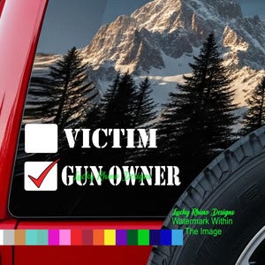 Pode incluir: Um decalque de vinil branco com as palavras "VICTIM" e "GUN OWNER" em negrito. O texto "GUN OWNER" tem uma marca de seleção ao lado. O fundo mostra uma cena de montanha com árvores.